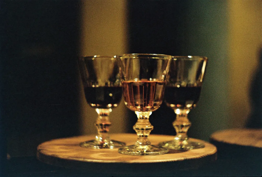 Three small stemmed glasses of amber and red wine on a round wooden board in low light