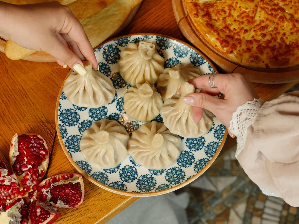 Two hands lifting khinkali by their knots from a blue patterned plate, khachapuri and pomegranate beside it
