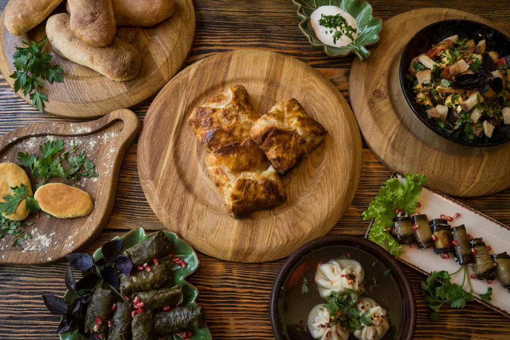 Wooden boards seen from above with bread, pastries, a salad, aubergine rolls, vine-leaf dolma and a bowl of khinkali in broth